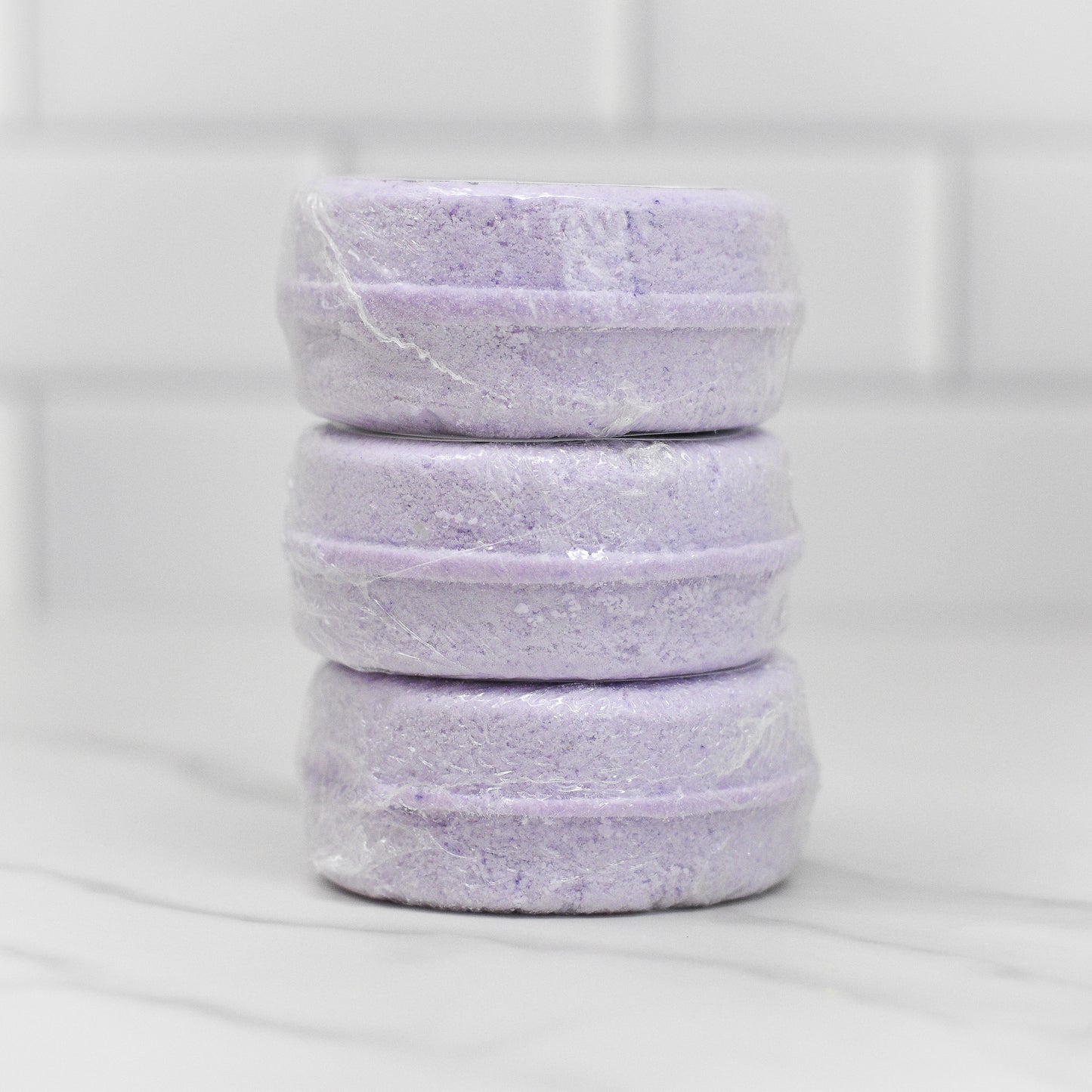Three stacked, lavender-colored Relax Shower Steamer bars from 570 Soap Co., wrapped in clear plastic, promise a calming shower experience as they sit against a white tiled background.