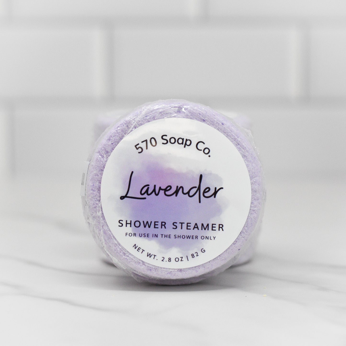 The Relax Shower Steamer - Lavender by 570 Soap Co. offers a calming shower experience. Wrapped in clear plastic and placed on a marble surface, its label reads "For use in the shower only" with a net weight of 2.8 oz / 82 g. A tiled wall in the background adds to the serene atmosphere.