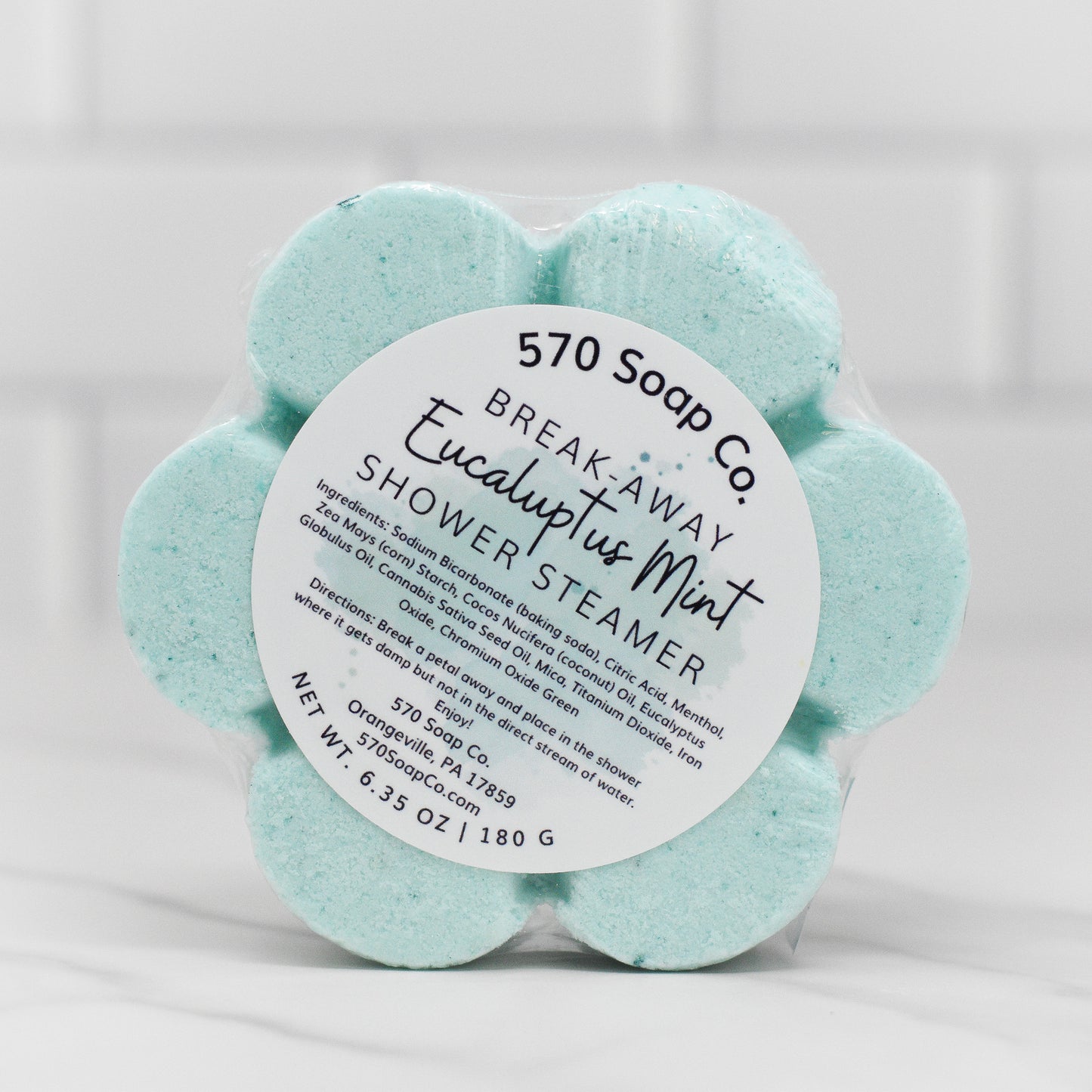 The Breathe Easy Shower Steamer in Eucalyptus Mint from 570 Soap Co. offers an aromatic experience. These flower-shaped steamers are encased in clear packaging, featuring a label that lists the ingredients on a white background, and have a net weight of 6.35 oz (180 g).