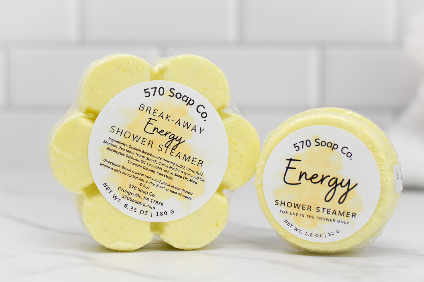 Two round yellow shower steamer tablets from 570 Soap Co. are showcased, each infused with invigorating citrus essential oils. One tablet is labeled as the Break-Away Energy Shower Steamer, while the other is referred to as the Energy Shower Steamer - Citrus. Both feature floral designs and are set against a pristine white tiled background.