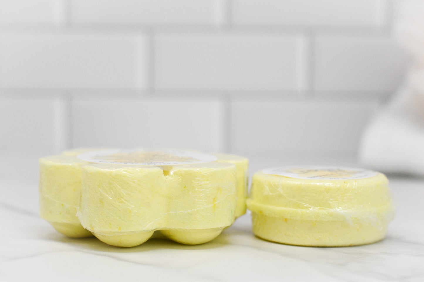 Two cylindrical yellow bars of the Energy Shower Steamer - Citrus by 570 Soap Co., infused with invigorating citrus essential oils, are wrapped in clear plastic and placed on a white marble surface. The steamer on the left is larger with a scalloped edge, set against a tiled white wall backdrop.