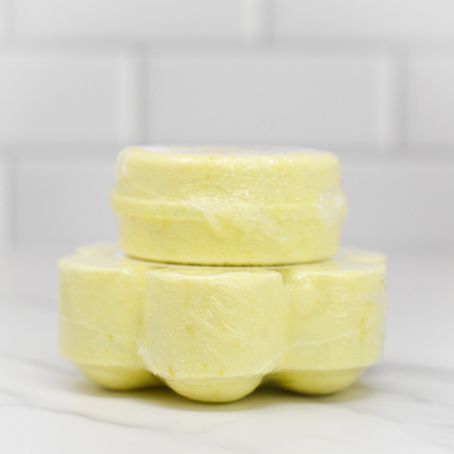 A duo of Energy Shower Steamers by 570 Soap Co. sits on a white surface, set against a backdrop of white tiled walls. Wrapped in clear plastic, these steamers are infused with citrus essential oils and feature a pale yellow hue.