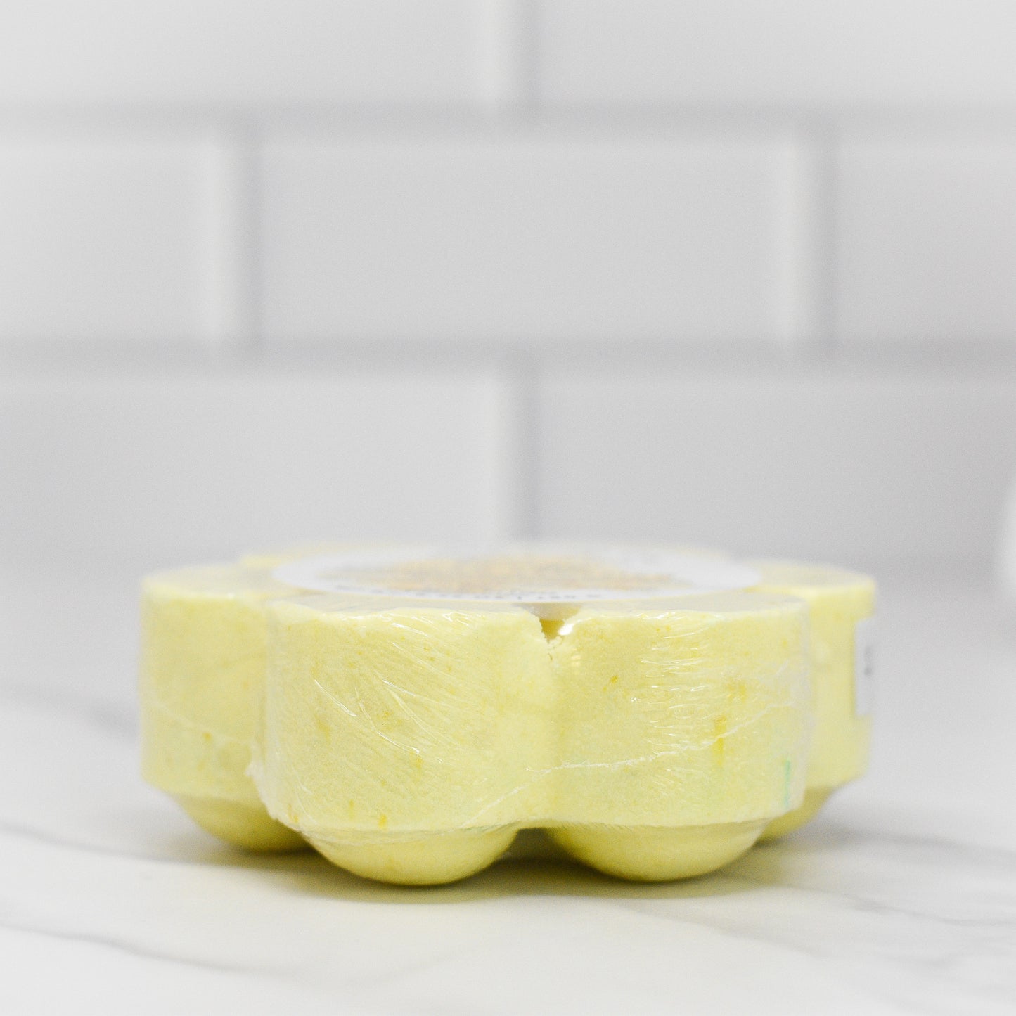 A close-up of a pack of six Energy Shower Steamer - Citrus from 570 Soap Co., featuring round yellow discs infused with citrus aromas, arranged in a circular shape on a white marble surface with a blurred white tiled background.