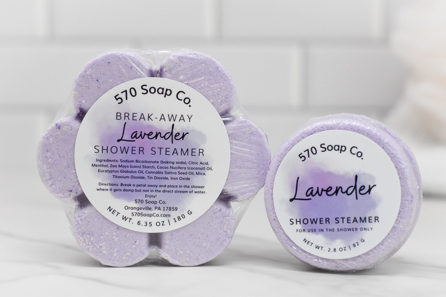 The image features two Relax Shower Steamer - Lavender products from 570 Soap Co. One is a packaged set of six flower-shaped pieces, while the other is a single round steamer. Ideal for creating a calming shower experience, the packaging includes ingredients and instructions for use.