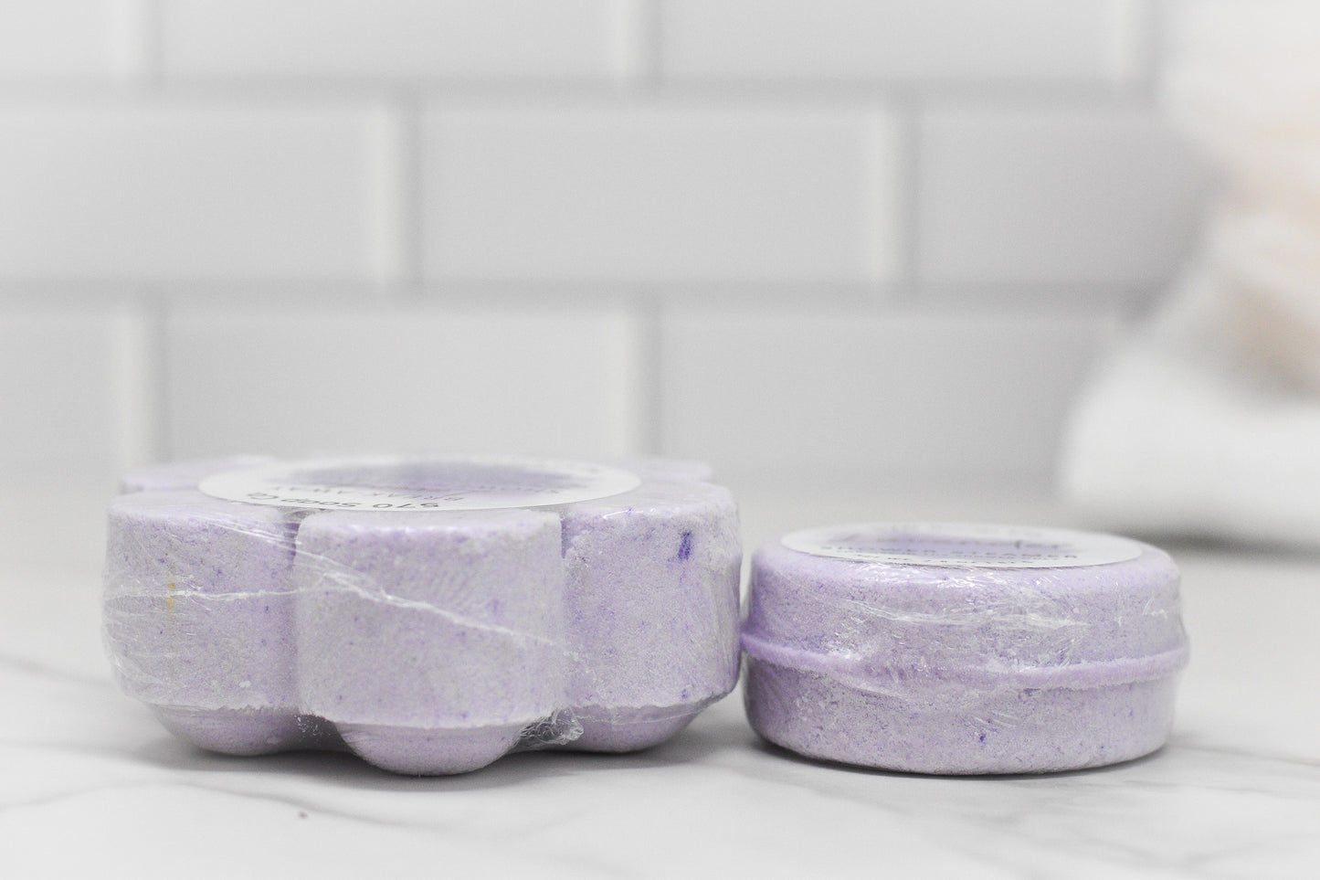 Two lavender-colored bath bombs are displayed on a marble surface in front of white subway tiles. The flower-shaped one on the left is partially wrapped in plastic, hinting at a calming shower experience with the Relax Shower Steamer - Lavender by 570 Soap Co., while the round one on the right promises tranquility.