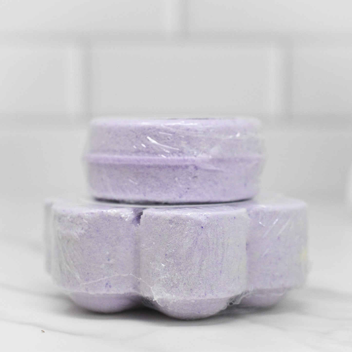 A stack of Relax Shower Steamer - Lavender rounds from 570 Soap Co., wrapped in plastic, sits on a marble countertop, promising a calming shower experience with soothing lavender and invigorating menthol aromas against the backdrop of a white tiled wall.