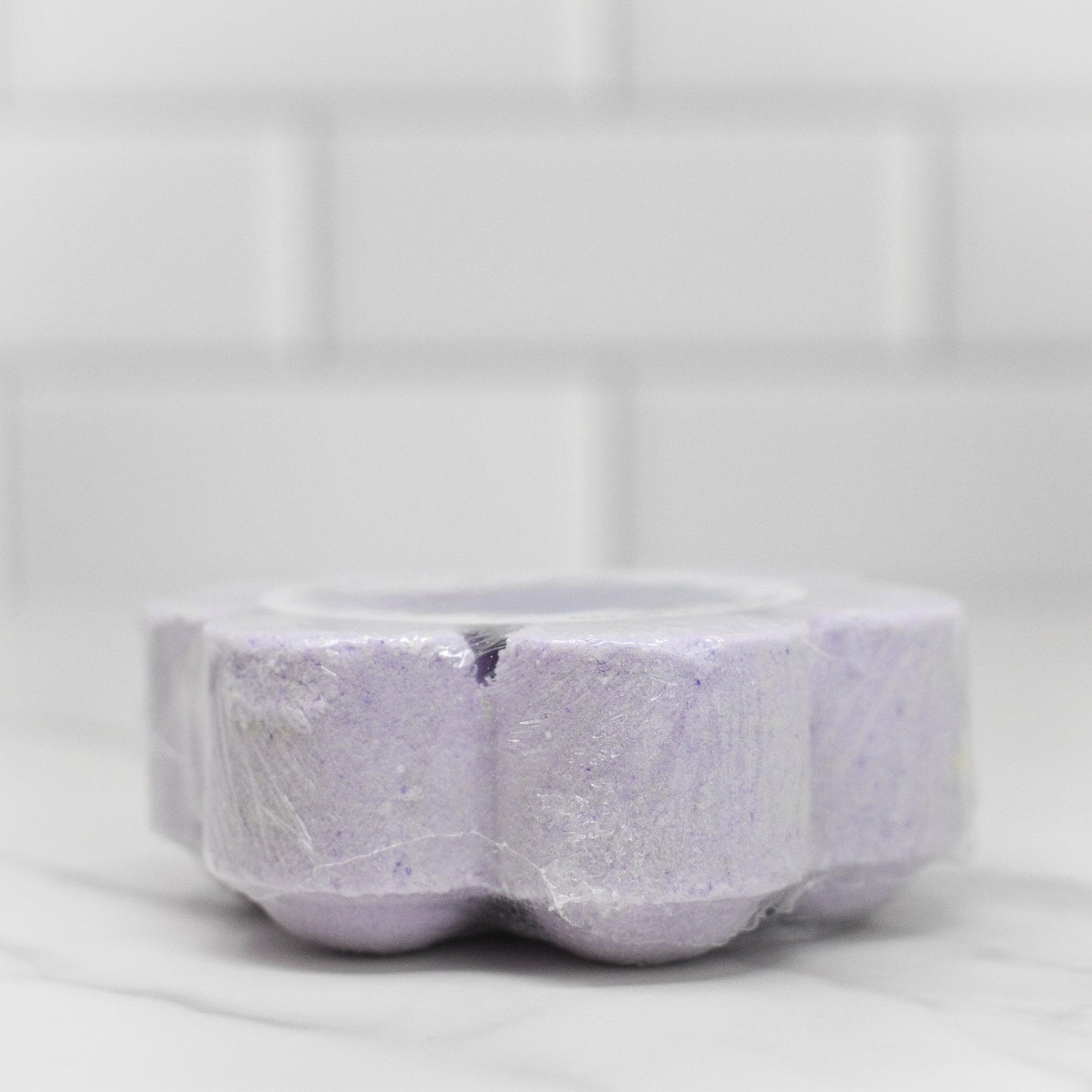 A Relax Shower Steamer - Lavender by 570 Soap Co., resembling a flower with petal-like sections, rests on a marble countertop beside a white tiled wall. This calming shower experience promises to transform your routine into pure relaxation. Wrapped in plastic, it's the perfect touch for serenity seekers.