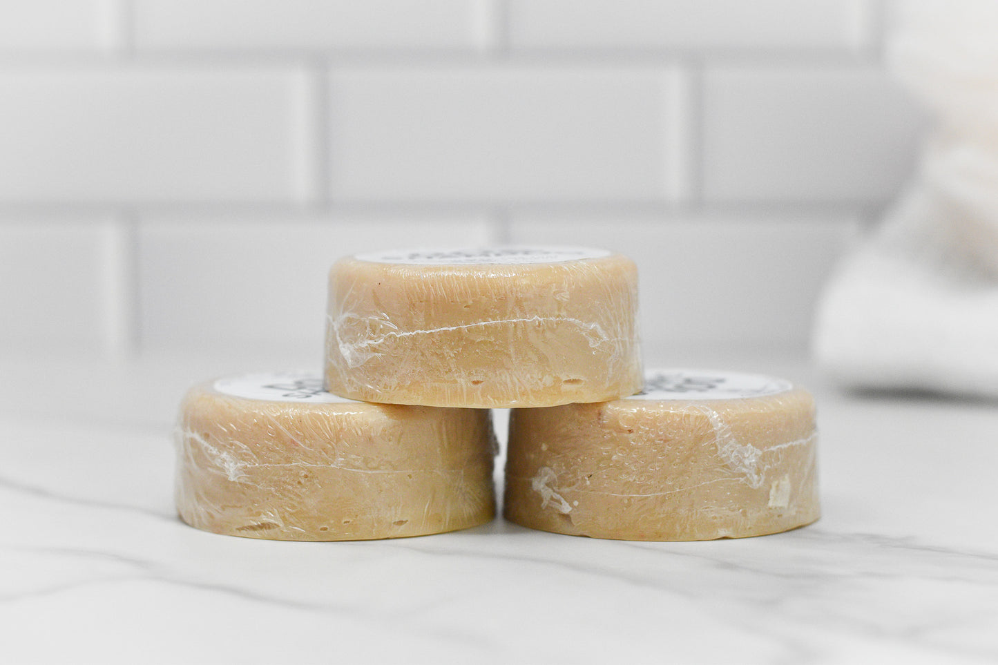 Three round bars of Dog Shampoo Bar - All Natural from 570 Soap Co., wrapped in plastic, are stacked on a white marble surface against a white tiled background. Infused with a soothing lavender scent, the shampoo bar boasts a light beige color, perfect for gentle cleansing.