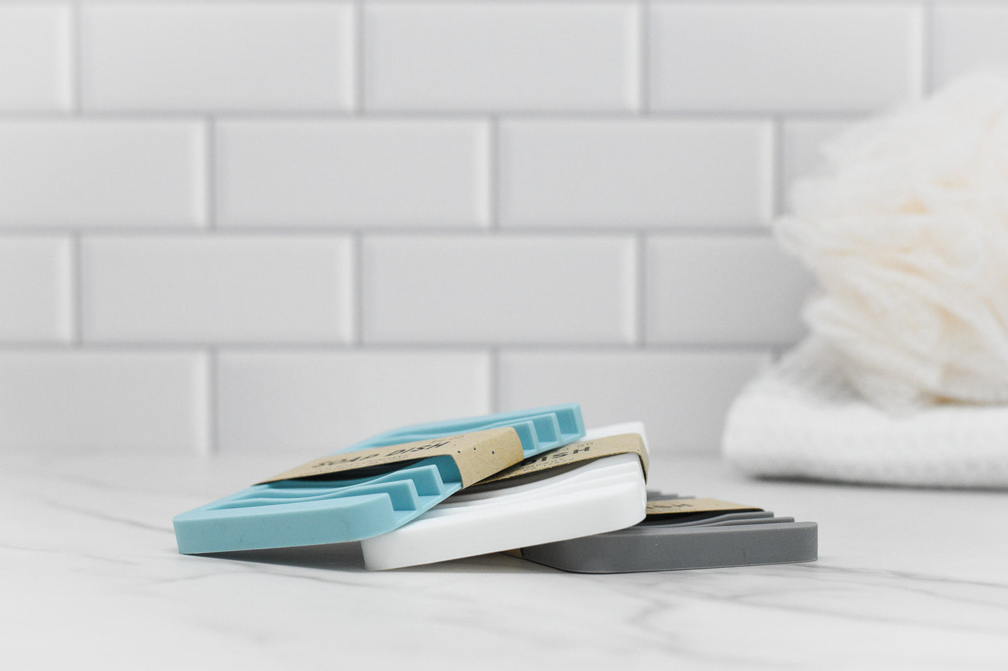 The 570 Soap Co. Soap Saver Silicone Draining Dishes, in blue, white, and gray, are beautifully stacked on a marble surface against a backdrop of white tiles. These self-draining designs enhance the longevity of your soap bar. In the background, a blurred white bath sponge can be seen.