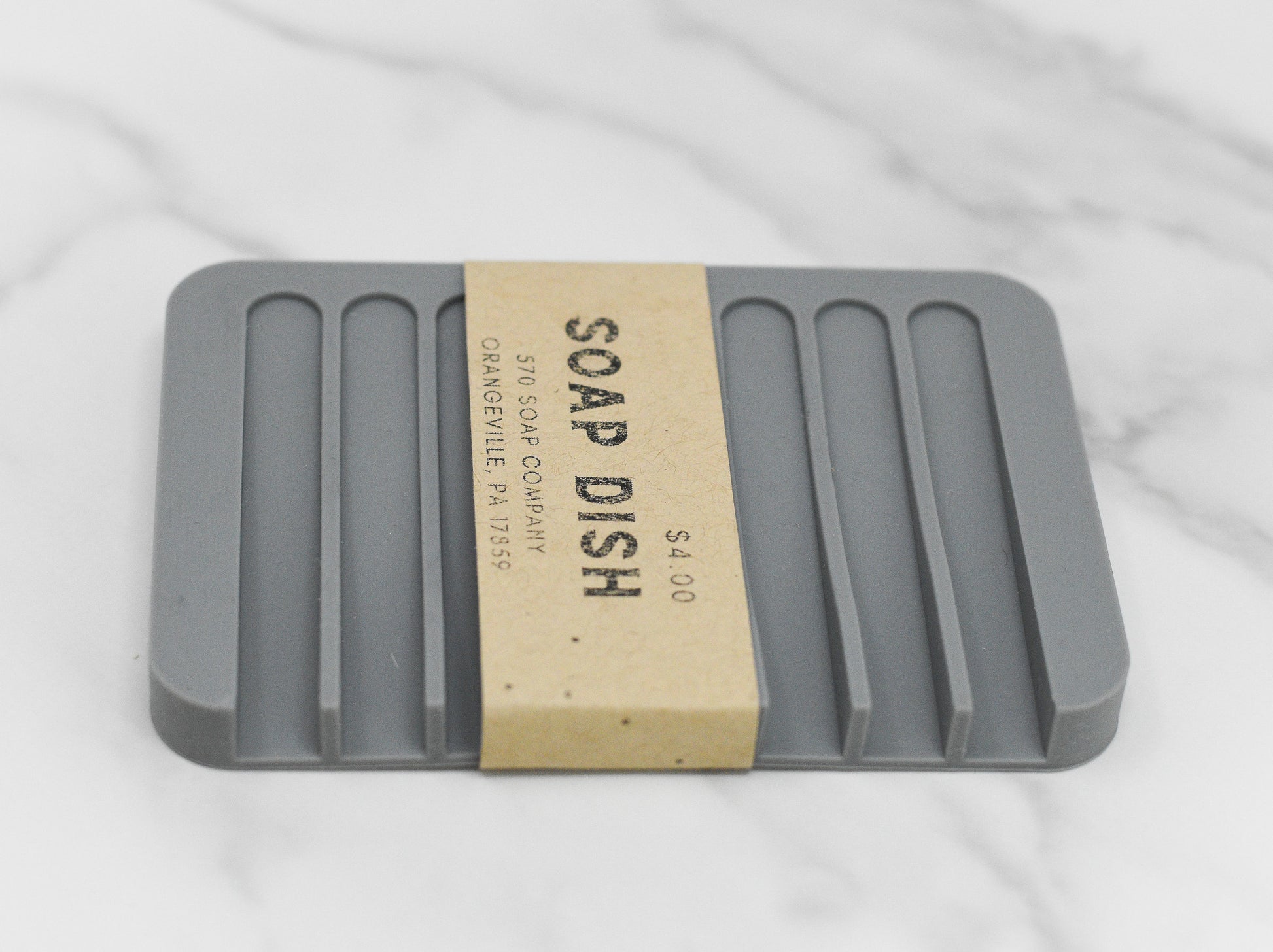 A gray, rectangular Soap Saver Silicone Draining Dish from 570 Soap Co. rests on a marble surface, featuring raised ridges for optimal drainage. Enveloped around the dish is a brown paper label that reads "Soap Saver Silicone Draining Dish," highlighting its design that promotes enhanced soap bar longevity through self-drainage.