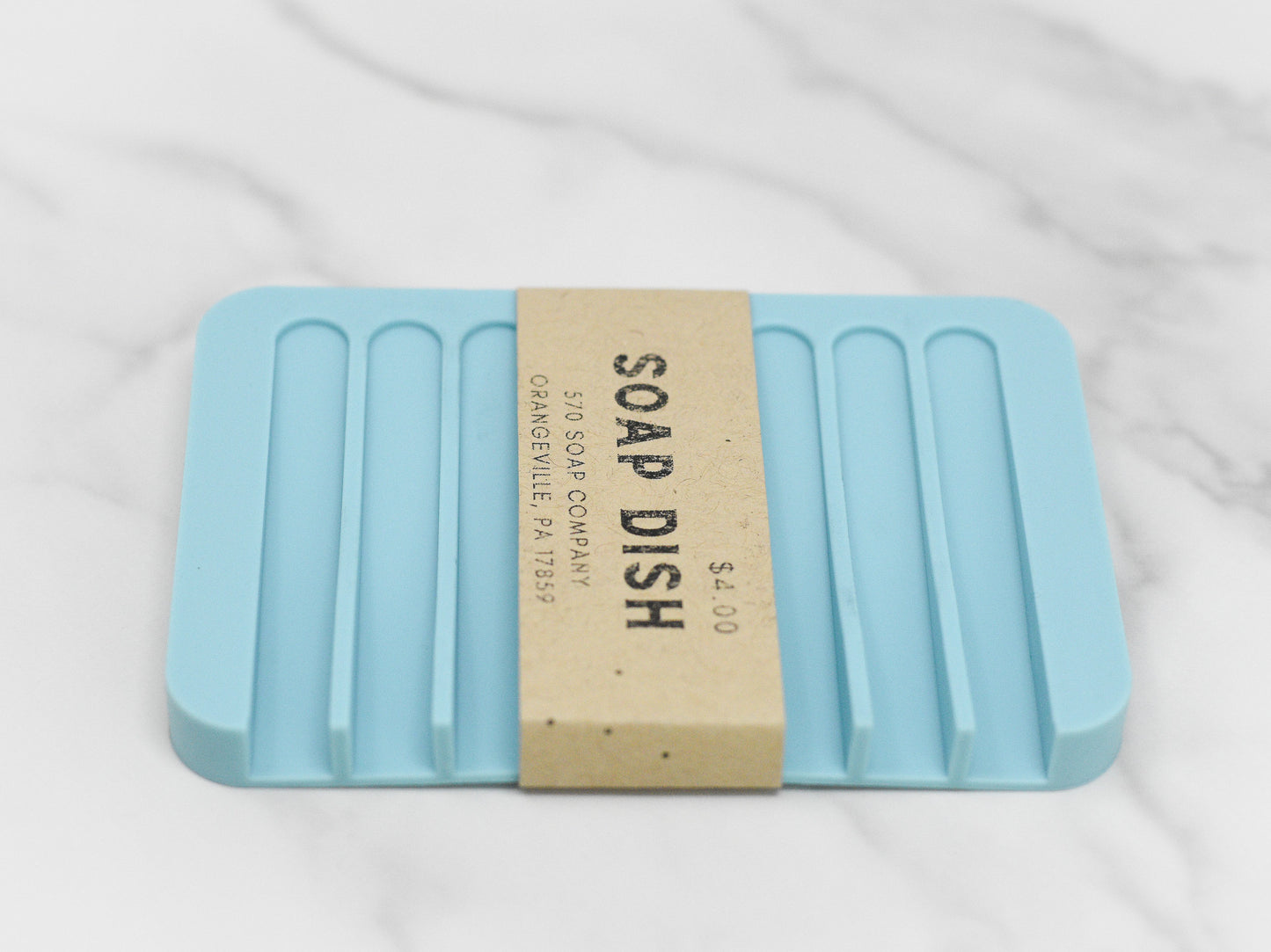 A light blue Soap Saver Silicone Draining Dish with grooves rests on a marble surface. A cardboard sleeve encircles it, displaying "570 Soap Co.," along with pricing and address details, designed to enhance soap bar longevity.