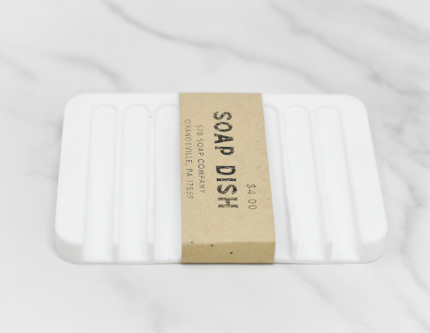A white silicone Soap Saver Silicone Draining Dish from 570 Soap Co. is placed on a marble surface. Enclosed in a brown paper band labeled with its name and priced at $4.00, this self-draining accessory is designed to extend the life of your soap bar. The band also includes an address for the company located in Pennsylvania.