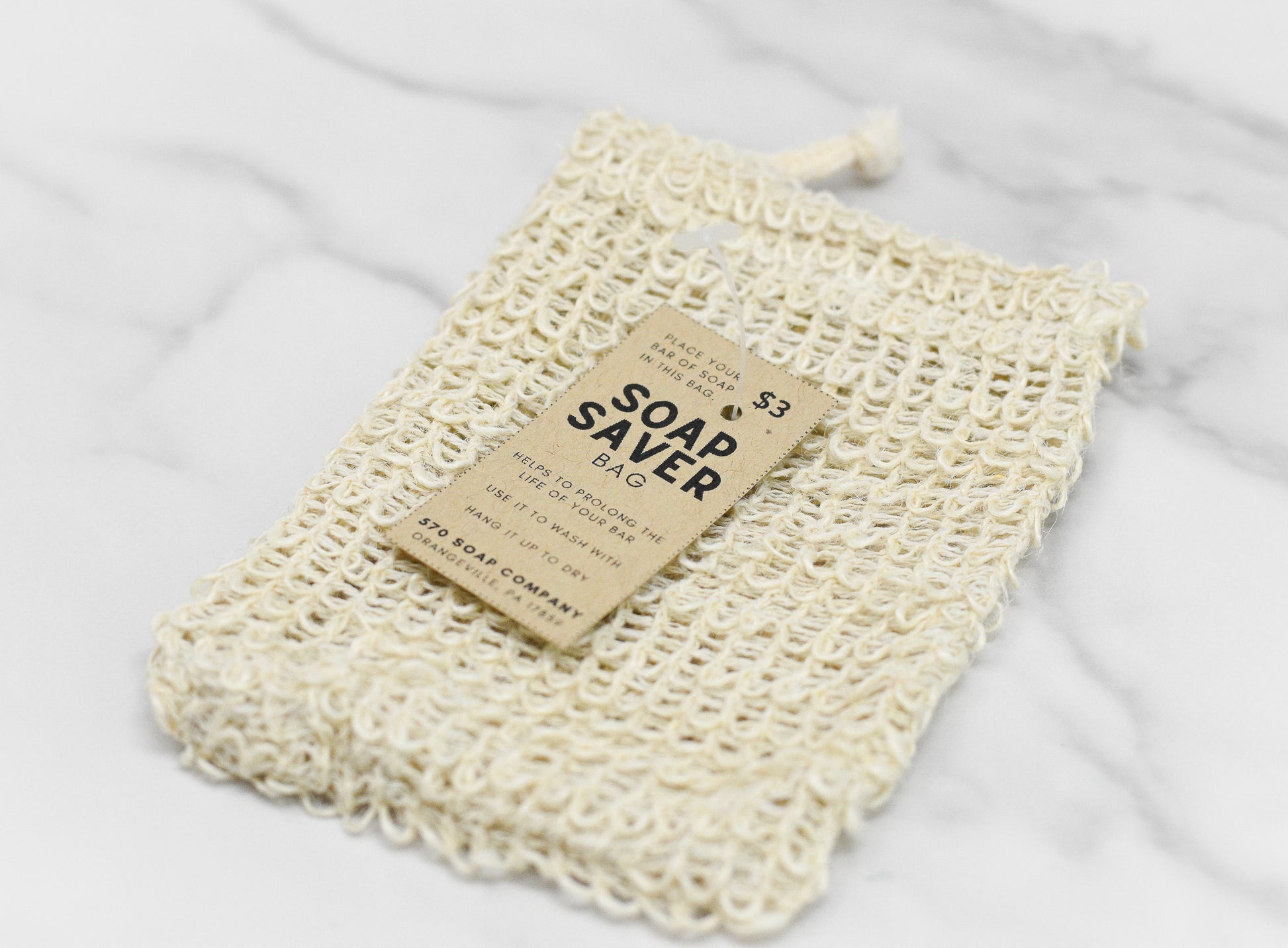 A beige Drawstring Soap Saving Mesh Bag from 570 Soap Co., crafted from sisal fibers, sits on a marble surface with a cardboard tag labeled "Drawstring Soap Saving Mesh Bag." The tag explains its gentle exfoliating benefits and provides usage instructions, priced at $3.