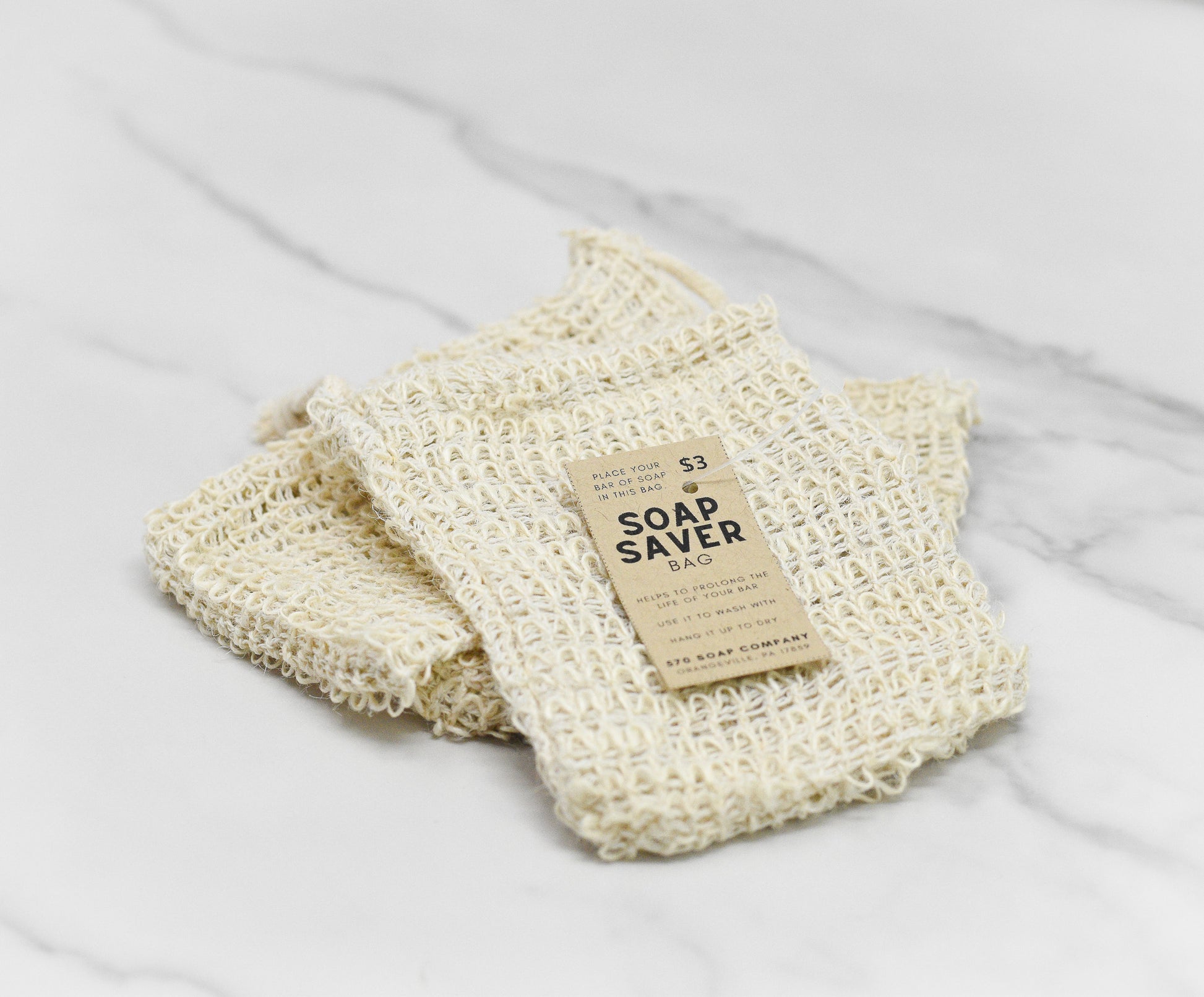 A cream-colored Drawstring Soap Saving Mesh Bag by 570 Soap Co., crafted from sisal fibers, sits on a marble surface. The beige label emphasizes its benefits and price, showcasing how it naturally exfoliates the skin while protecting your favorite soaps.