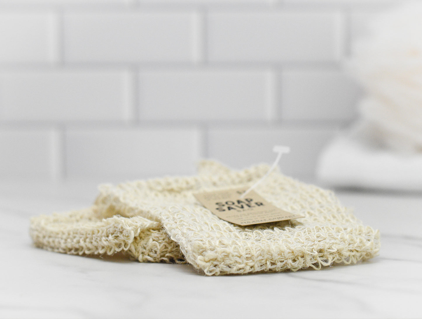 A beige, textured washcloth with a tag on top rests beside the 570 Soap Co.'s Drawstring Soap Saving Mesh Bag on the white marble counter. Made from sisal fibers, this mesh bag gently exfoliates skin, while white subway tiles in the background enhance the clean and minimalist setting.
