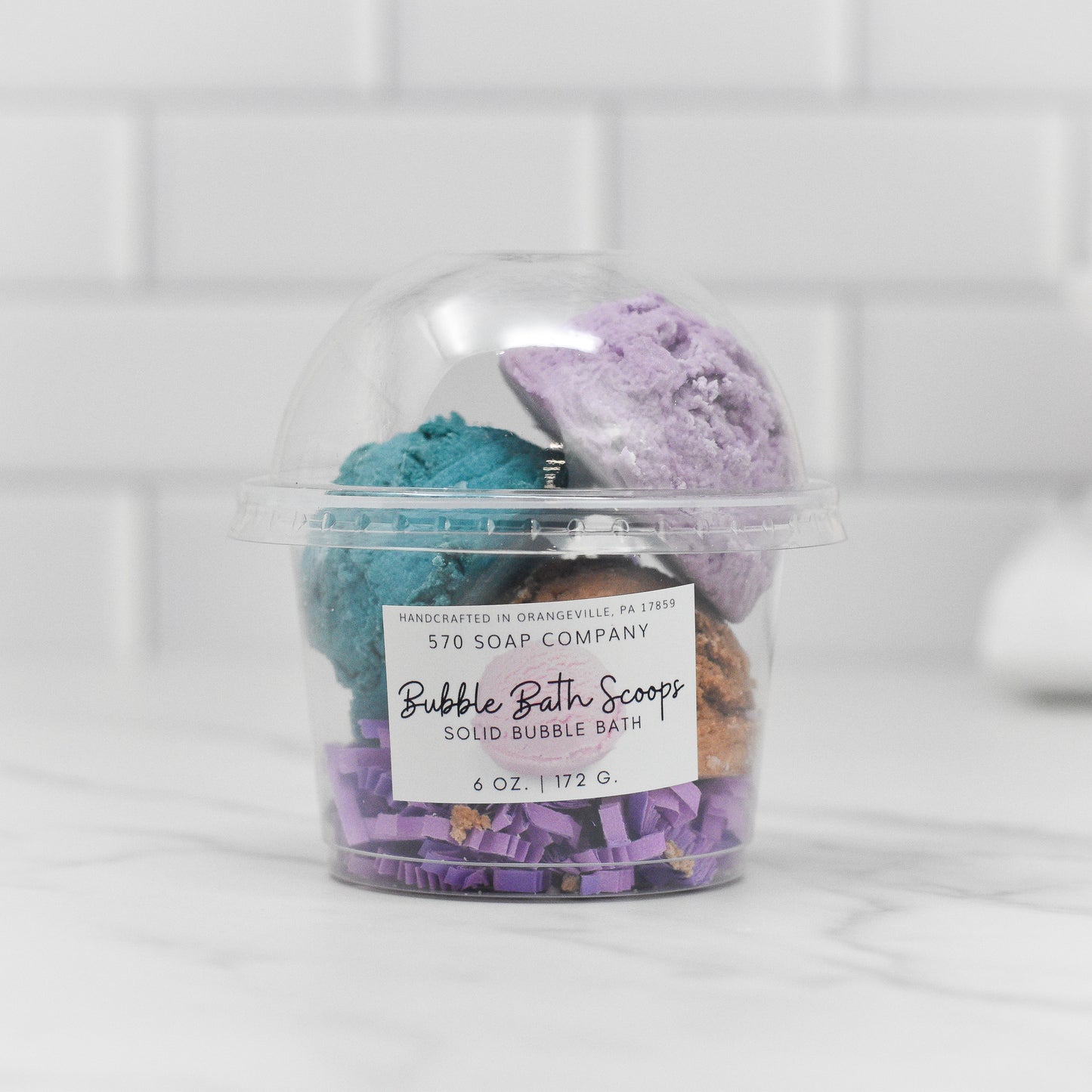 A clear container displays vibrant solid bubble bath scoops in shades of teal, orange, and purple. The label reads, "Solid Bubble Bath Scoop by 570 Soap Co." Set against a backdrop of white tiles, these charming scoops turn bath time into a soothing retreat for your skin.