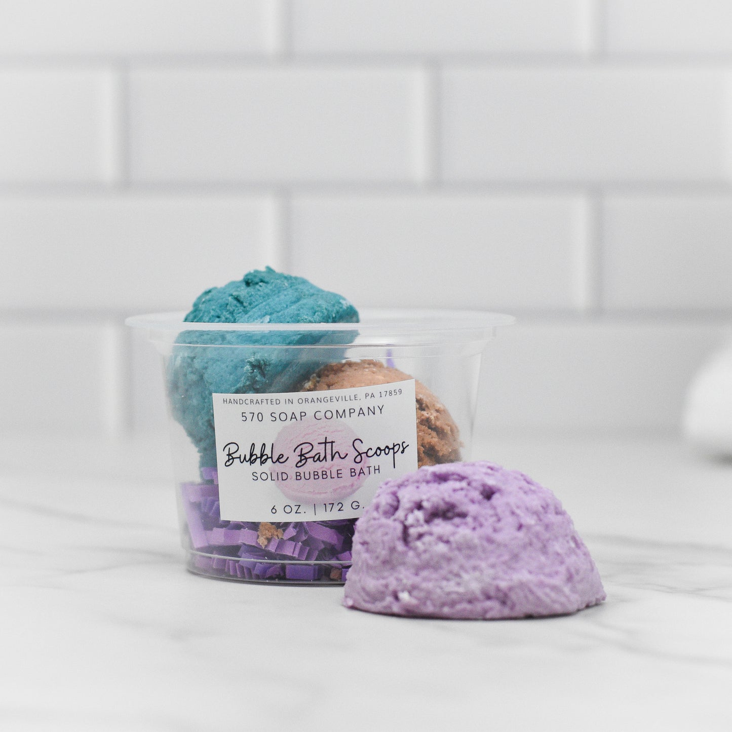 A transparent container labeled as Solid Bubble Bath Scoop houses three vibrant bath products from 570 Soap Co. resembling ice cream scoops in teal, brown, and purple hues. A purple scoop is set in front of the container, offering luxurious bubbles with its skin-nourishing formula, all arranged against a backdrop of white tiles.