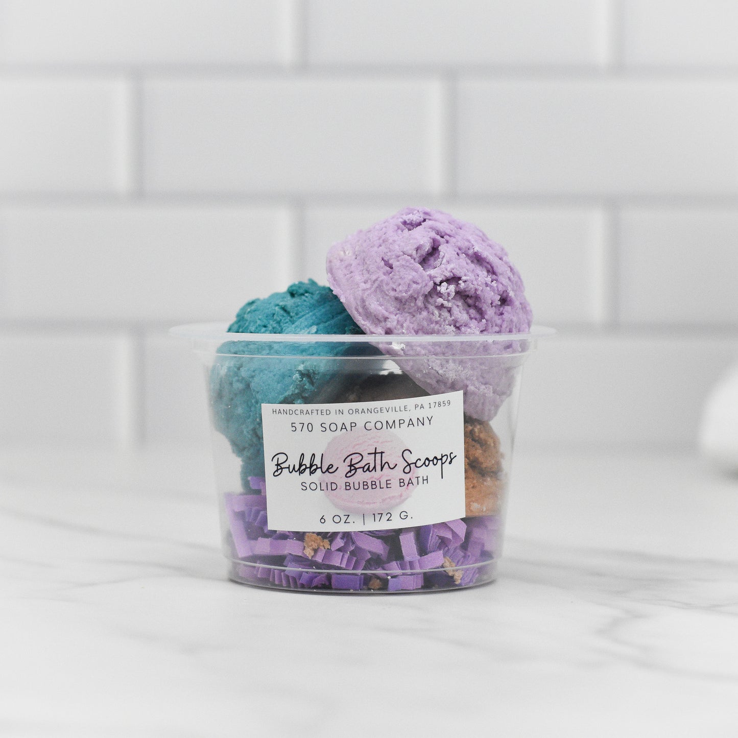 A clear plastic container holds two vibrant, scoop-shaped bubble bath solids—one in purple and the other in teal. The label reads Solid Bubble Bath Scoop by 570 Soap Co. These luxurious bubbles are displayed on a white marble surface against a backdrop of white tiled walls.