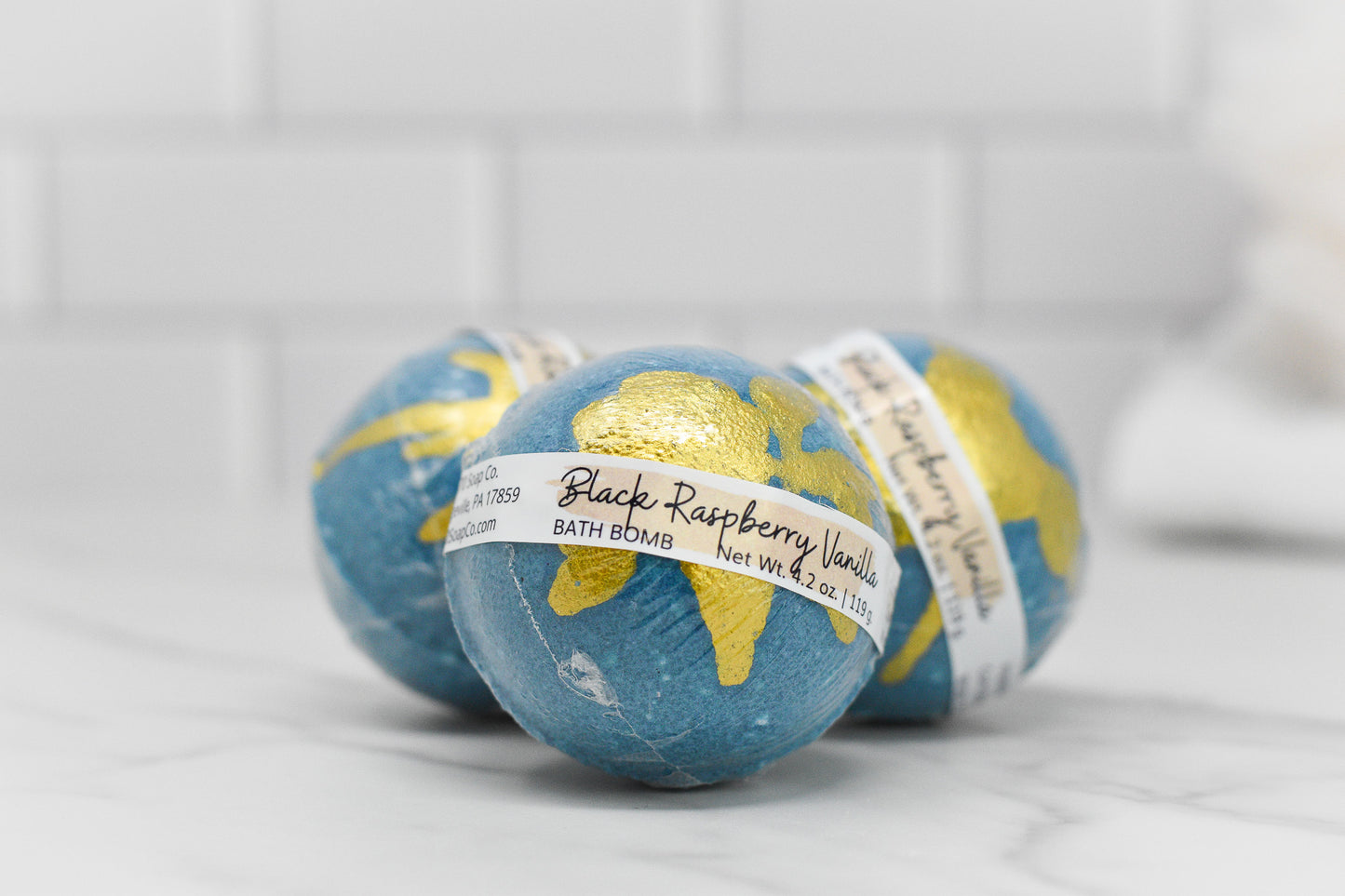 A trio of Black Raspberry Vanilla Teal Bath Bombs by 570 Soap Co., adorned with gold accents, offers a promise of relaxation. They are elegantly displayed against a white, tiled background on a marble surface.