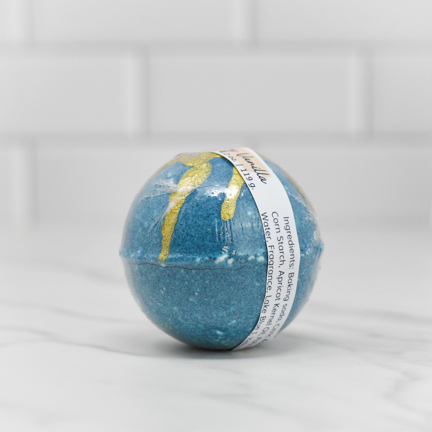 A Black Raspberry Vanilla Teal Bath Bomb from 570 Soap Co. rests on a white marble surface, radiating relaxation. Its spherical shape is accented with gold streaks and it features a label around the middle showing ingredients and other details. The backdrop is a white tiled wall, adding to the clean, minimalist aesthetic with subtle hints of its signature fragrance.