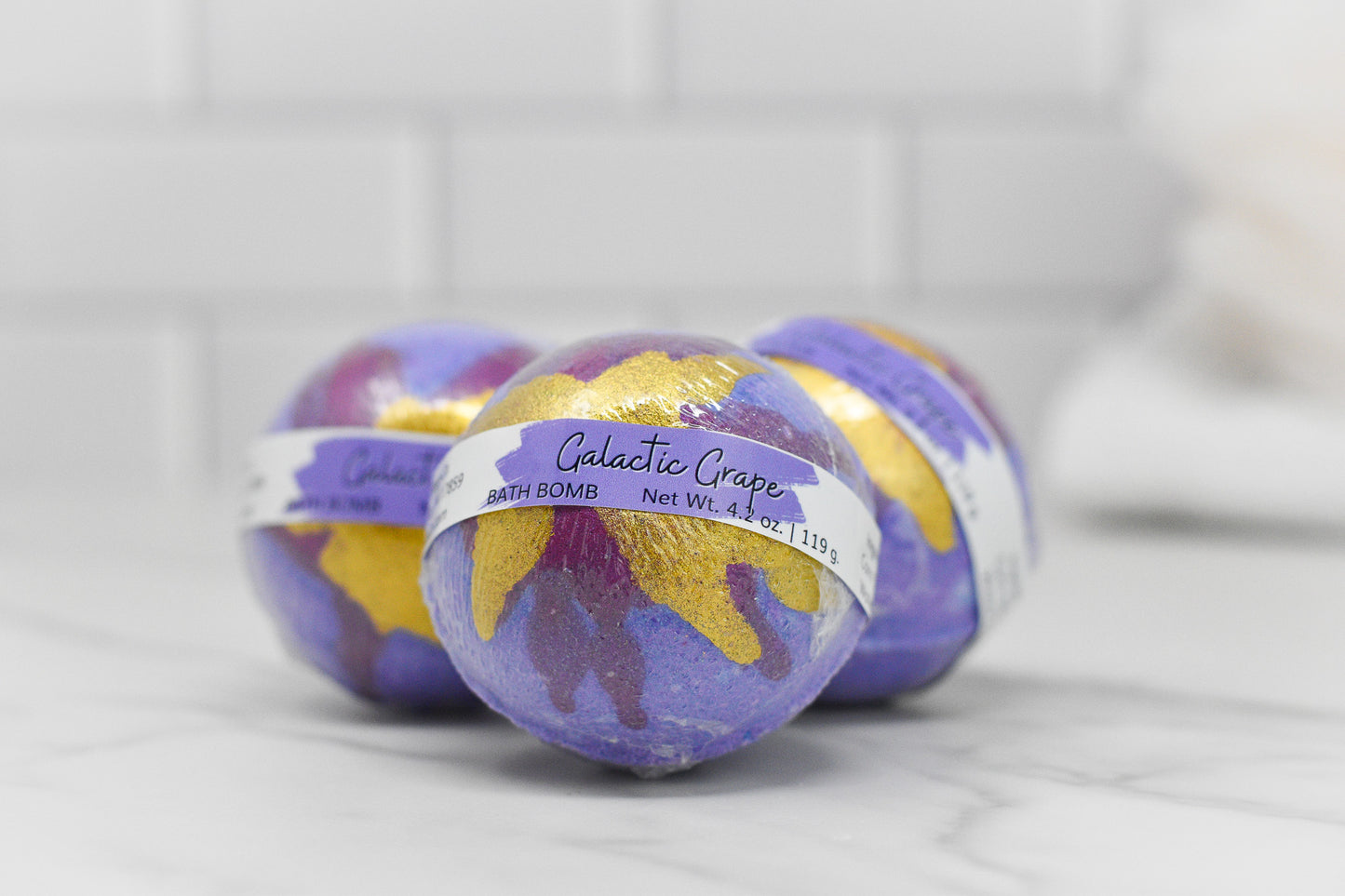 Three purple and gold Galactic Grape Bath Bomb: Round products by 570 Soap Co. are on display on a white marble surface, with a blurred white tiled background.