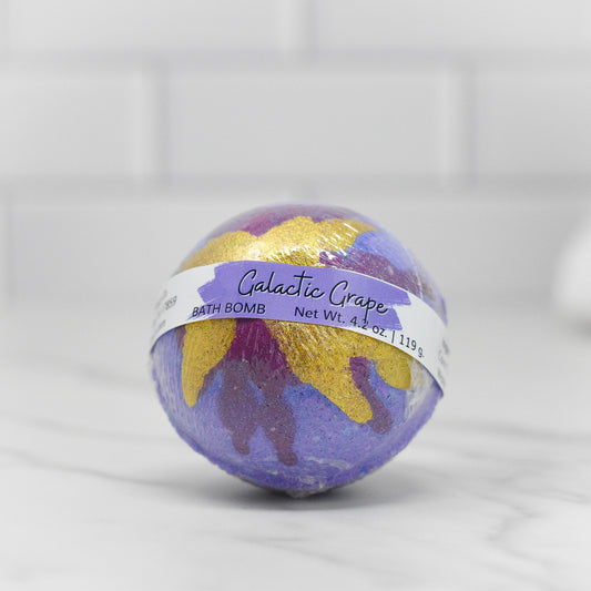 The 4.2 oz Galactic Grape Bath Bomb by 570 Soap Co., with gold and pink accents, offers a cosmic aroma that transports you to another world, resting elegantly on white marble against a blurred tiled backdrop for a nourishing skin experience.