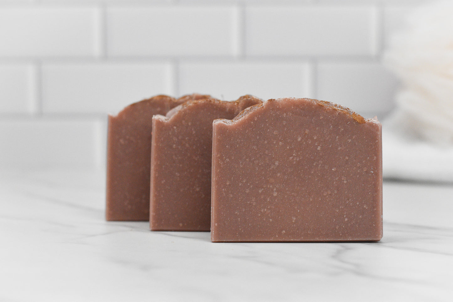Three bars of 570 Soap Co.'s Wild Earth Hunters Blend Bar Soap - Dirt Soap, featuring curved tops, are neatly arranged on a white marble surface. Infused with kaolin clay, they emit a unique dirt-scented aroma. The backdrop consists of white subway tiles, adding to the clean and minimalist aesthetic.