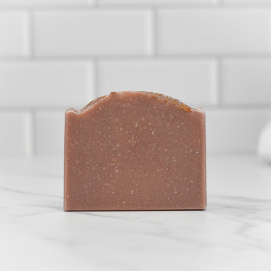 A bar of Wild Earth Hunters Blend Bar Soap - Dirt Soap by 570 Soap Co., featuring a smooth, reddish-brown surface, is artfully positioned on a white marble countertop with a backdrop of white tiled backsplash. The soap emits an intriguing dirt-scented fragrance, ideal for those who embrace rustic charm.