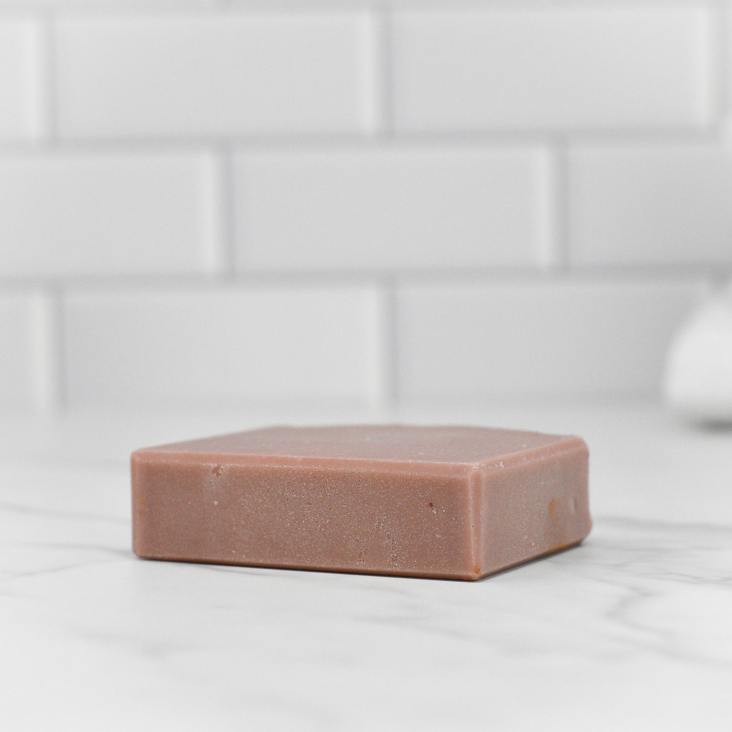 A single Wild Earth Hunters Blend Bar Soap - Dirt Soap from 570 Soap Co. sits on a white marble surface, its smooth texture and rectangular shape enhanced by the white tiled wall behind it. Enriched with kaolin clay and infused with a dirt-scented fragrance, it exudes an earthy aroma while providing nourishing benefits.