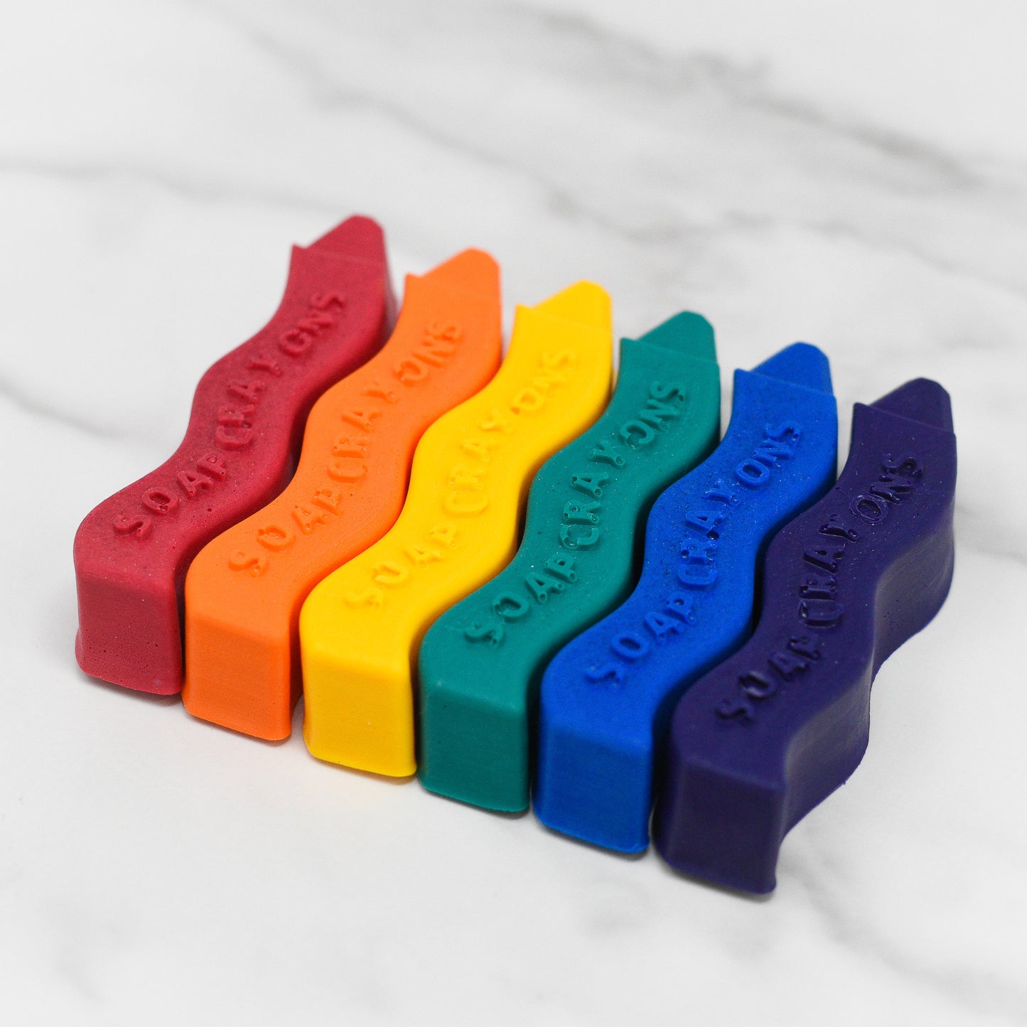 Six kid-friendly soaps, designed in the shape of crayons, rest neatly on a marble surface in vibrant shades of red, orange, yellow, green, blue, and purple. Each soap is embossed with "Unleash Creativity Soap Crayons," by 570 Soap Co., adding a splash of color and fun to bath time.
