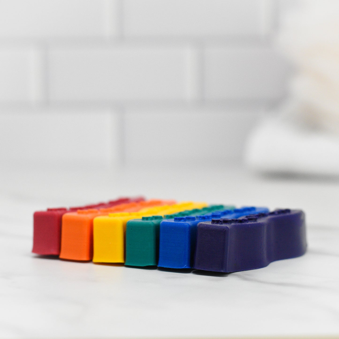 A row of vibrant Unleash Creativity Soap Crayons by 570 Soap Co., ideal for a kid-friendly bath time experience, is arranged in rainbow order on a white marble surface, with a white tiled wall in the background.