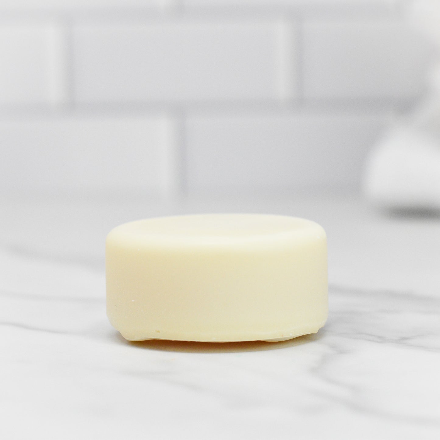 A small, round Zesty Lemon Dish Soap Bar by 570 Soap Co., creamy white with a subtle lemon scent, sits on a smooth white marble surface in an eco-friendly kitchen. The blurred white tile background enhances its all-natural appeal.