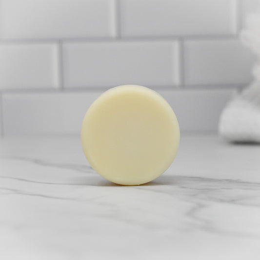 A round, pale yellow bar of Dish Soap Bar - All Natural by 570 Soap Co. sits on a marble countertop, with the backdrop showcasing white tile walls that enhance the clean and minimalist vibe of this eco-friendly scene.