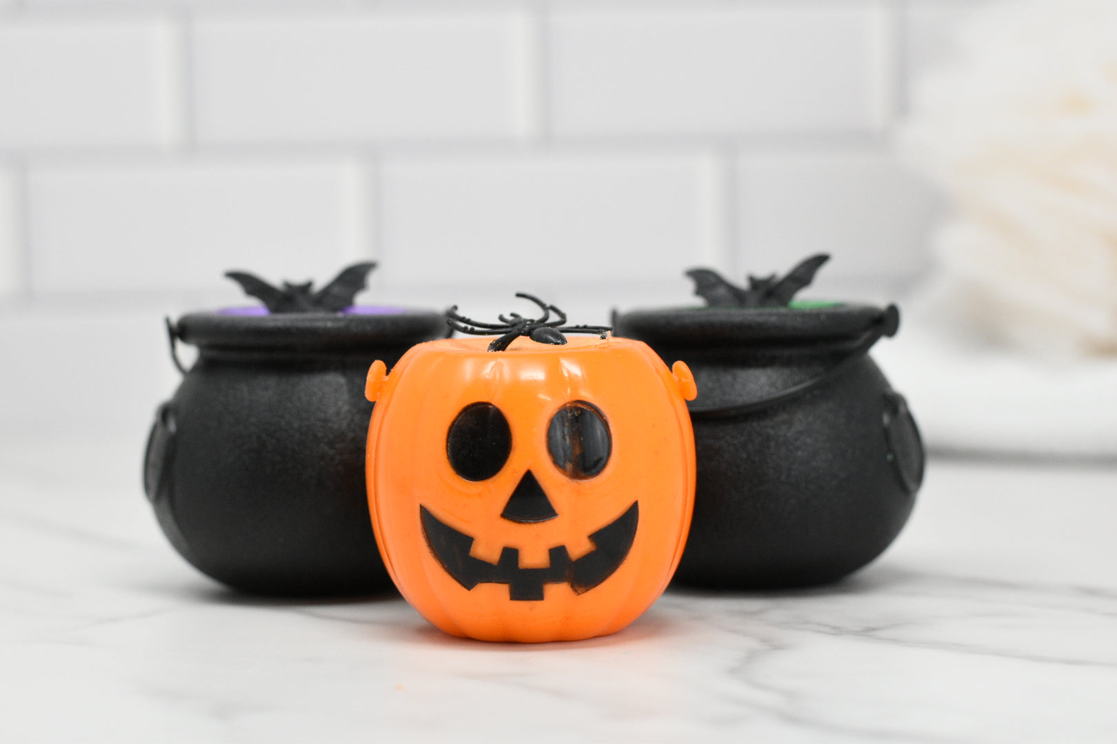 A delightful scene unfolds with a small orange pumpkin-shaped container featuring a smiling jack-o-lantern face, positioned in front of two enchanting black Witches Brew Bath Bomb Fizzing Cauldrons from 570 Soap Co. Adorned with bat decorations, these cauldrons invite you to imagine the magic of a witch's bubbling concoction. The entire setup is beautifully contrasted against a white tiled background, creating the perfect setting for an enchanting bath experience.