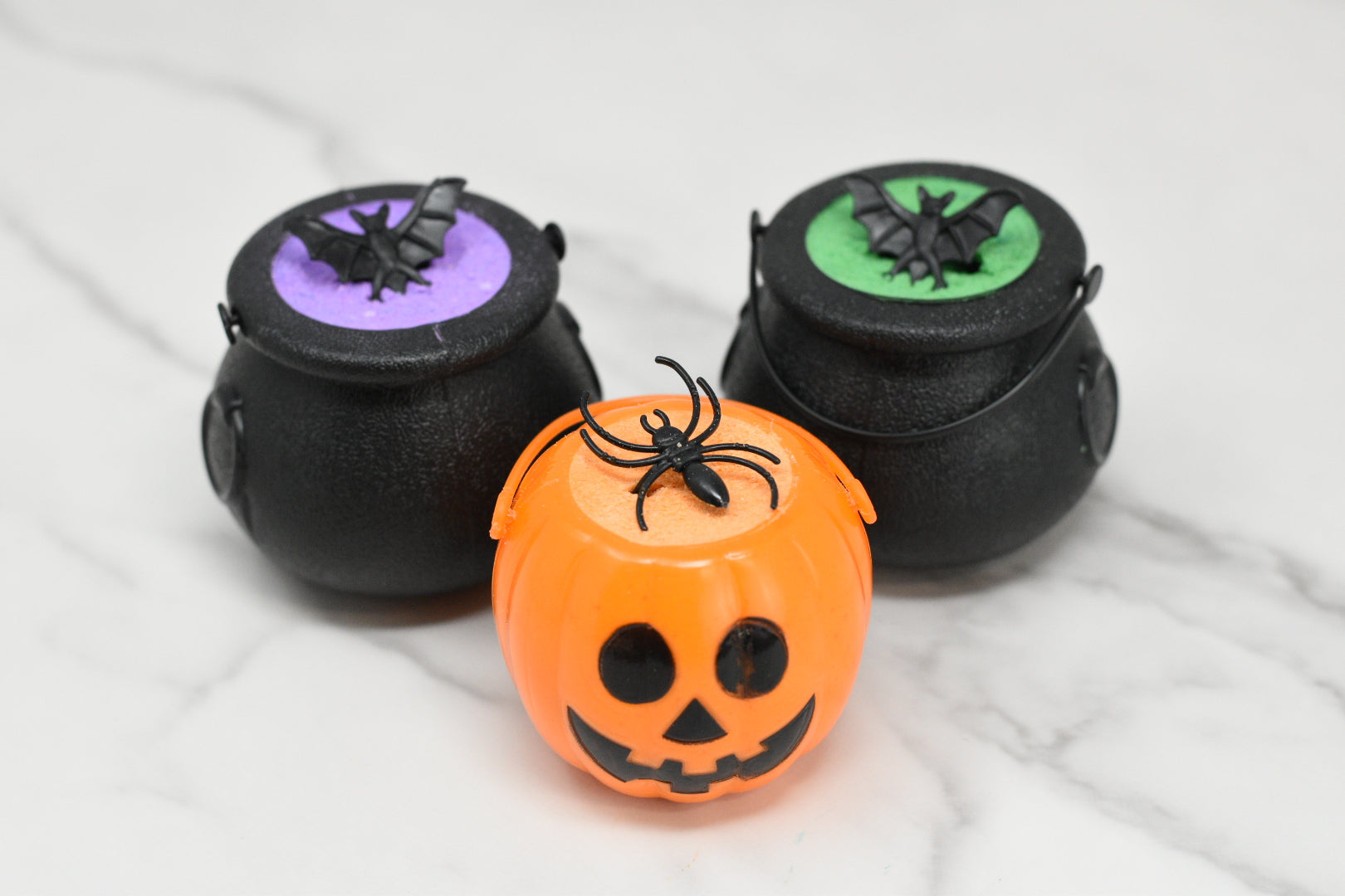 On a marble surface, there are three small Halloween-themed containers from 570 Soap Co. At the front is an orange pumpkin adorned with a black spider, and behind it sit two black cauldrons known as Witches Brew Bath Bomb Fizzing Cauldrons, each featuring vibrant purple and green tops with bats, promising an enchanting bath experience reminiscent of a witch’s bubbling concoction.