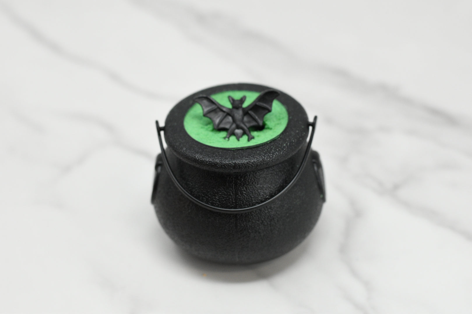 A small black cauldron with a handle, adorned with a green circle and black bat design on its lid, sits on a marble surface, inviting you to an enchanting bath experience. Aptly named the Witches Brew Bath Bomb Fizzing Cauldron from 570 Soap Co., it promises bewitched relaxation with its hidden bath bomb magic.