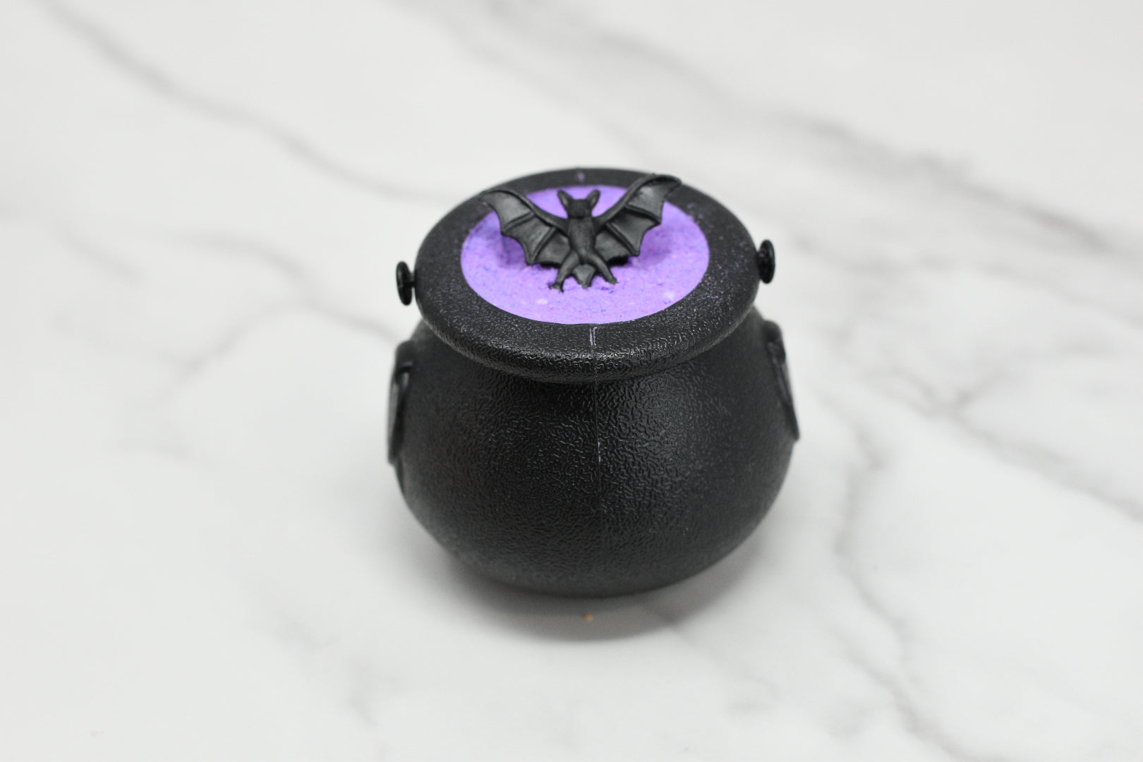 The Witches Brew Bath Bomb Fizzing Cauldron by 570 Soap Co. offers an enchanting bath experience with its black cauldron-shaped design on a marble surface. Filled with purple powder and crowned with a black bat decoration, it transforms your soak into magic as it dissolves into a shimmering bath bomb.
