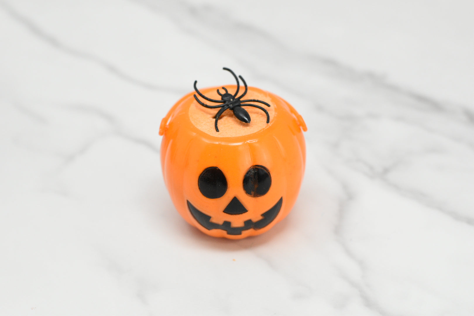 A Witches Brew Bath Bomb Fizzing Cauldron from 570 Soap Co., featuring an orange pumpkin-shaped container with a black smiling face and a faux black spider on top, sits on a white marbled surface, evoking the charm of a magical cauldron and promising an enchanting bath experience.