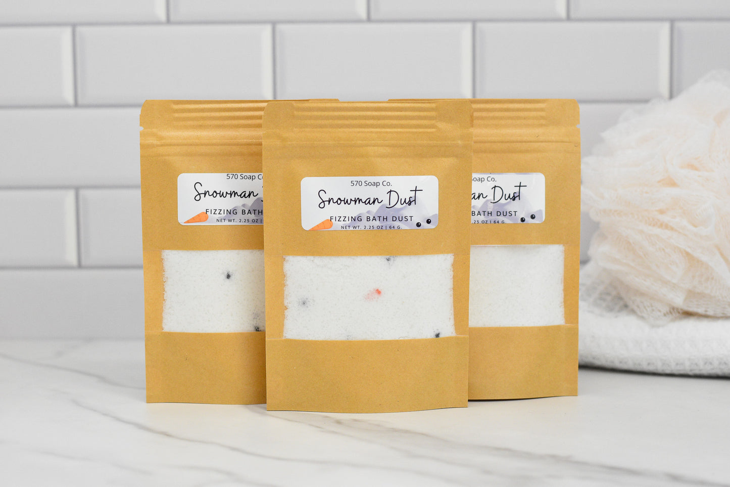Three kraft paper bags of Snowman Dust - Whimsical Fizzing Bath Elixir by 570 Soap Co., are arranged on a marble counter with a white tiled background. These are infused with Apricot Kernel Oil for a soothing experience. A white bath pouf is partially visible on the right.