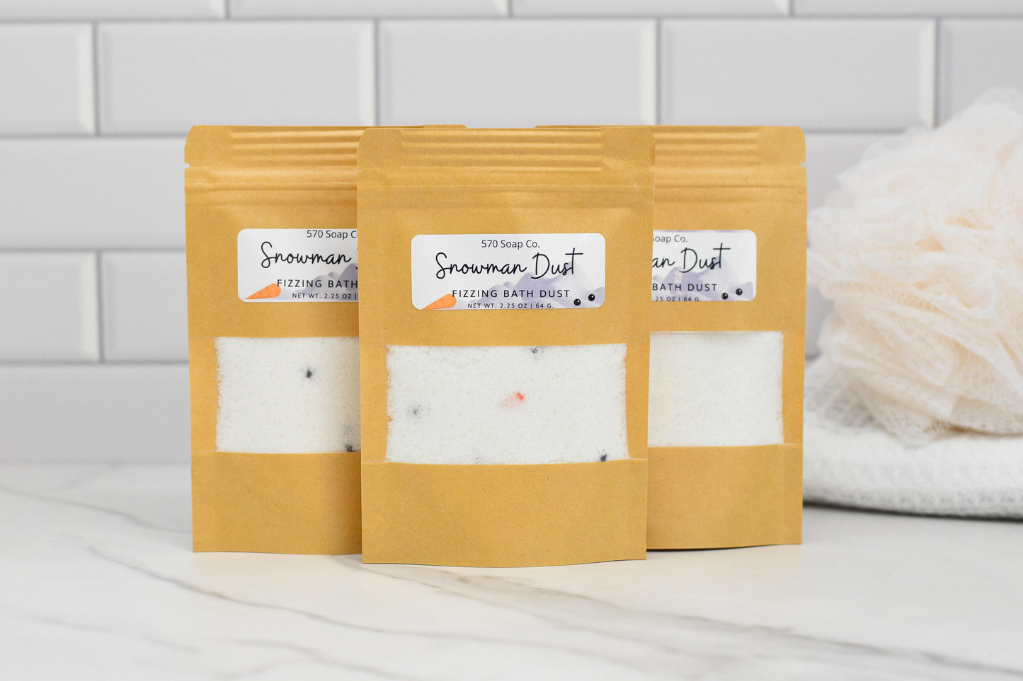 Three brown paper pouches of 570 Soap Co.'s Snowman Dust - Whimsical Fizzing Bath Elixir are arranged on a marble surface in front of a white tiled wall, prepared to transform your bath time. A white mesh bath sponge is partially visible on the right, enhancing the serene setting.