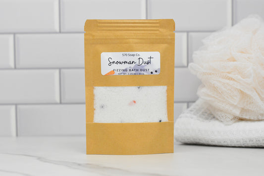 A kraft paper bag featuring the product Snowman Dust - Whimsical Fizzing Bath Elixir by 570 Soap Co. is placed on a white marble surface. In the background, white tiled walls and a plush bath sponge subtly suggest the infusion of apricot kernel oil inside.