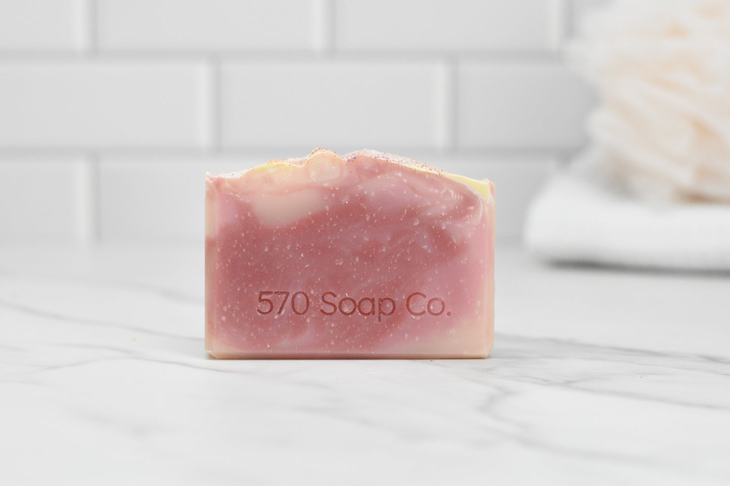 A Sweet Serenade Soap Bar by 570 Soap Co., featuring a pink and cream swirl and embossed with the brand's name, is displayed on a marble surface. It is set against a white tiled wall with a soft, blurred background that evokes a bathroom environment. This soap bar is crafted with natural ingredients to provide an exotic fragrance experience.