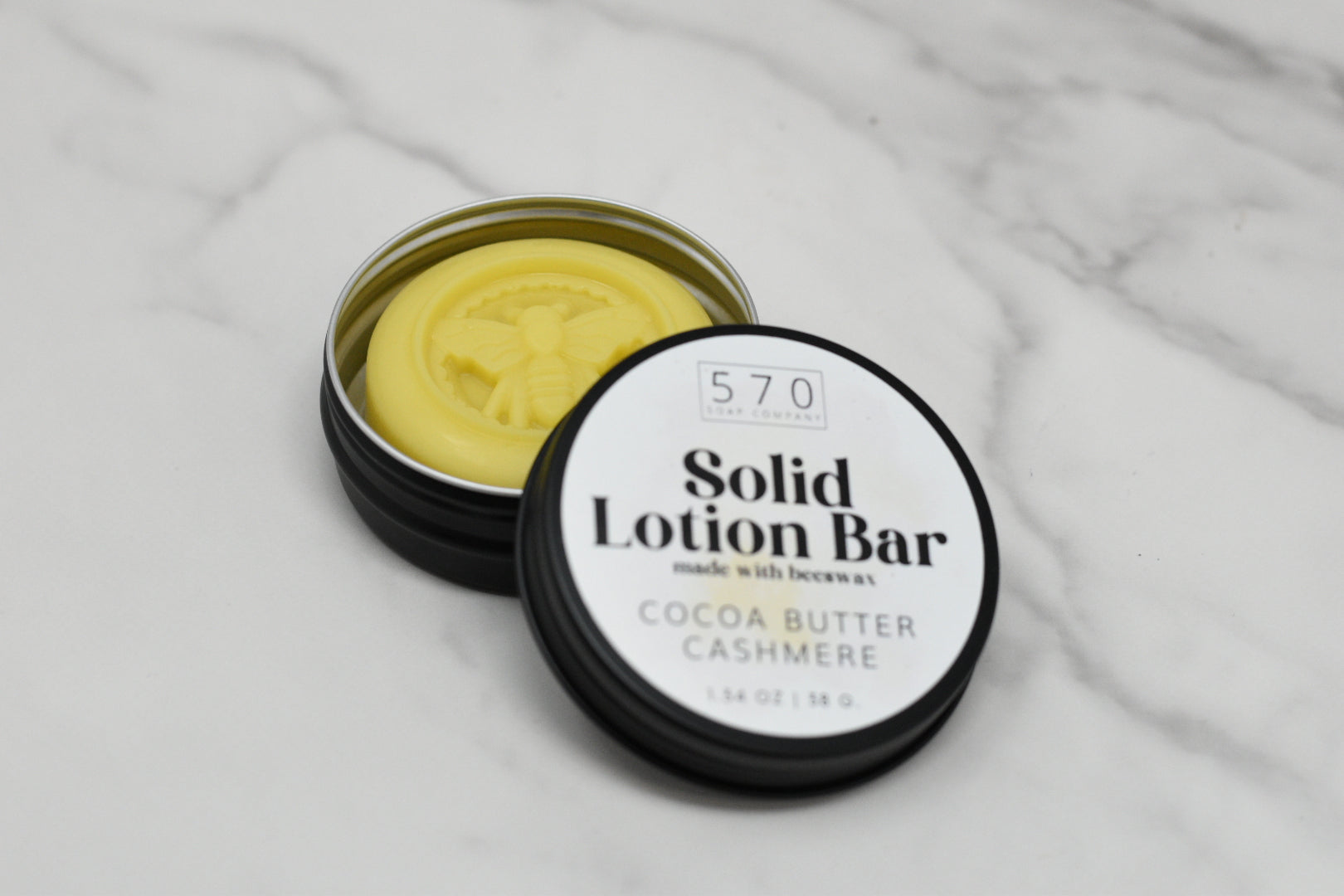 An open tin containing a yellow solid lotion bar engraved with a bee is placed on a marble surface. The adjacent lid features the text "Solid Lotion Bar - made with beeswax" by 570 Soap Co., emphasizing its function as a portable moisturizer crafted from natural ingredients, including Cocoa Butter Cashmere.
