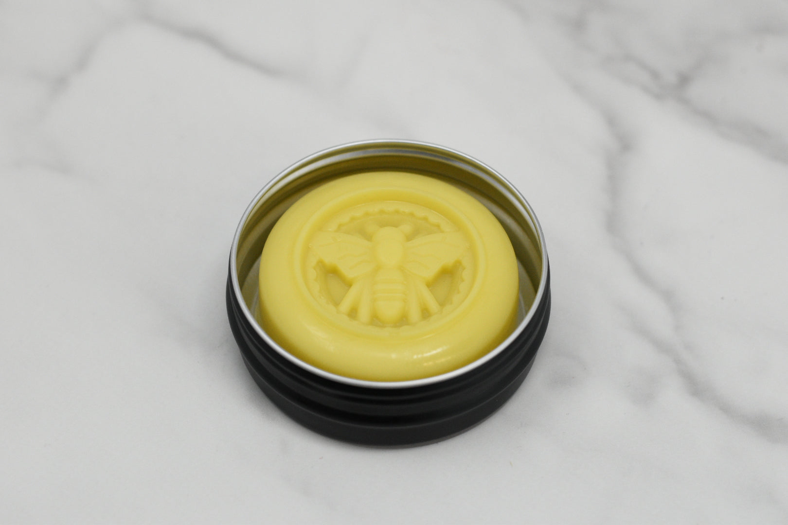 A round tin container from 570 Soap Co., showcasing a yellow balm with a raised bee design on the lid, rests elegantly on a marble surface. The Solid Lotion Bar - made with beeswax, is crafted with natural ingredients, offering an ideal portable moisturizer for hydration on the go.