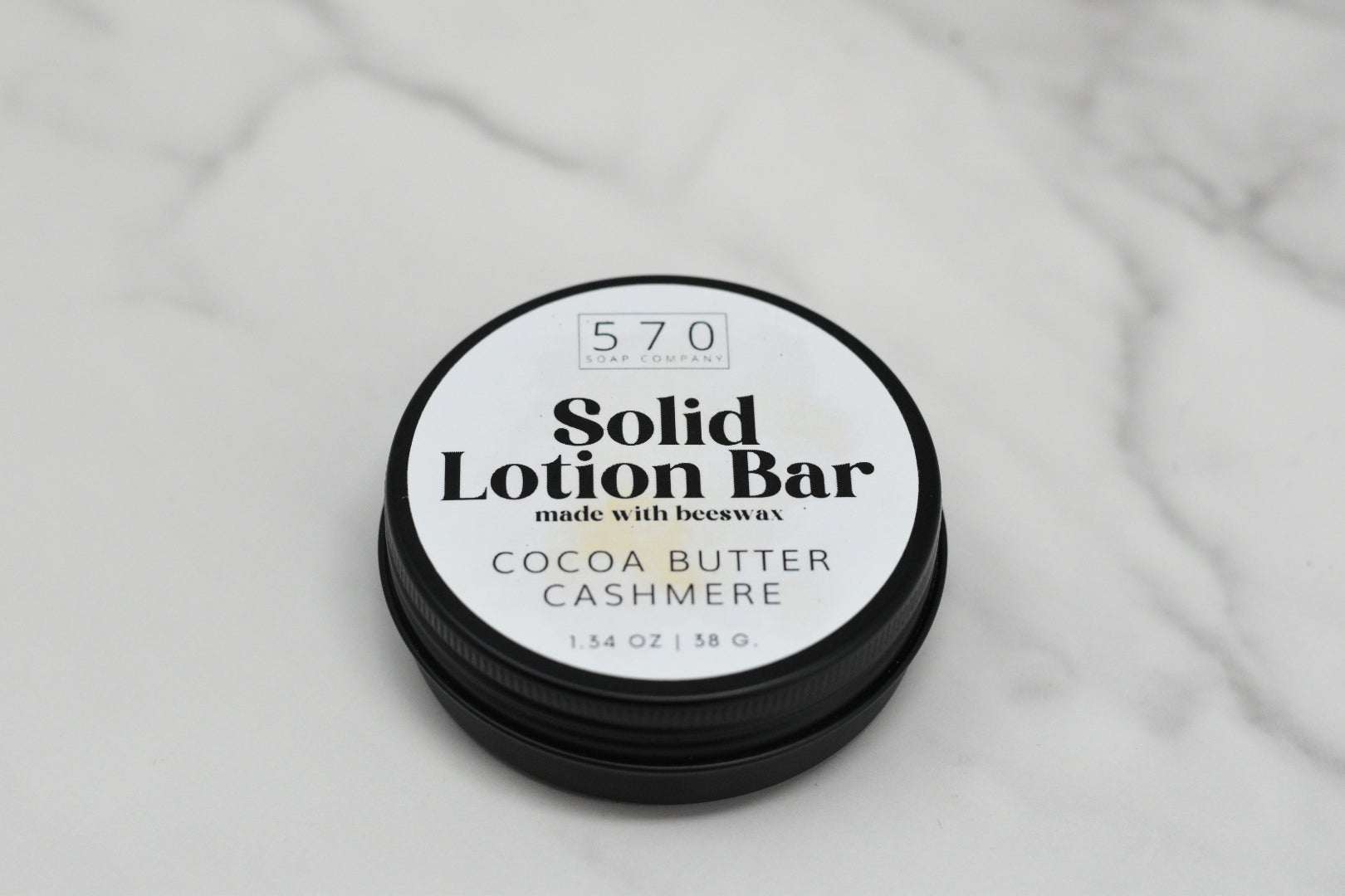 A circular container of Solid Lotion Bar, a handy moisturizer crafted with natural ingredients such as beeswax, rests on a marble surface. The label indicates "Cocoa Butter Cashmere" and 1.34 oz / 38 g. The brand name, 570 Soap Co., is elegantly displayed at the top.