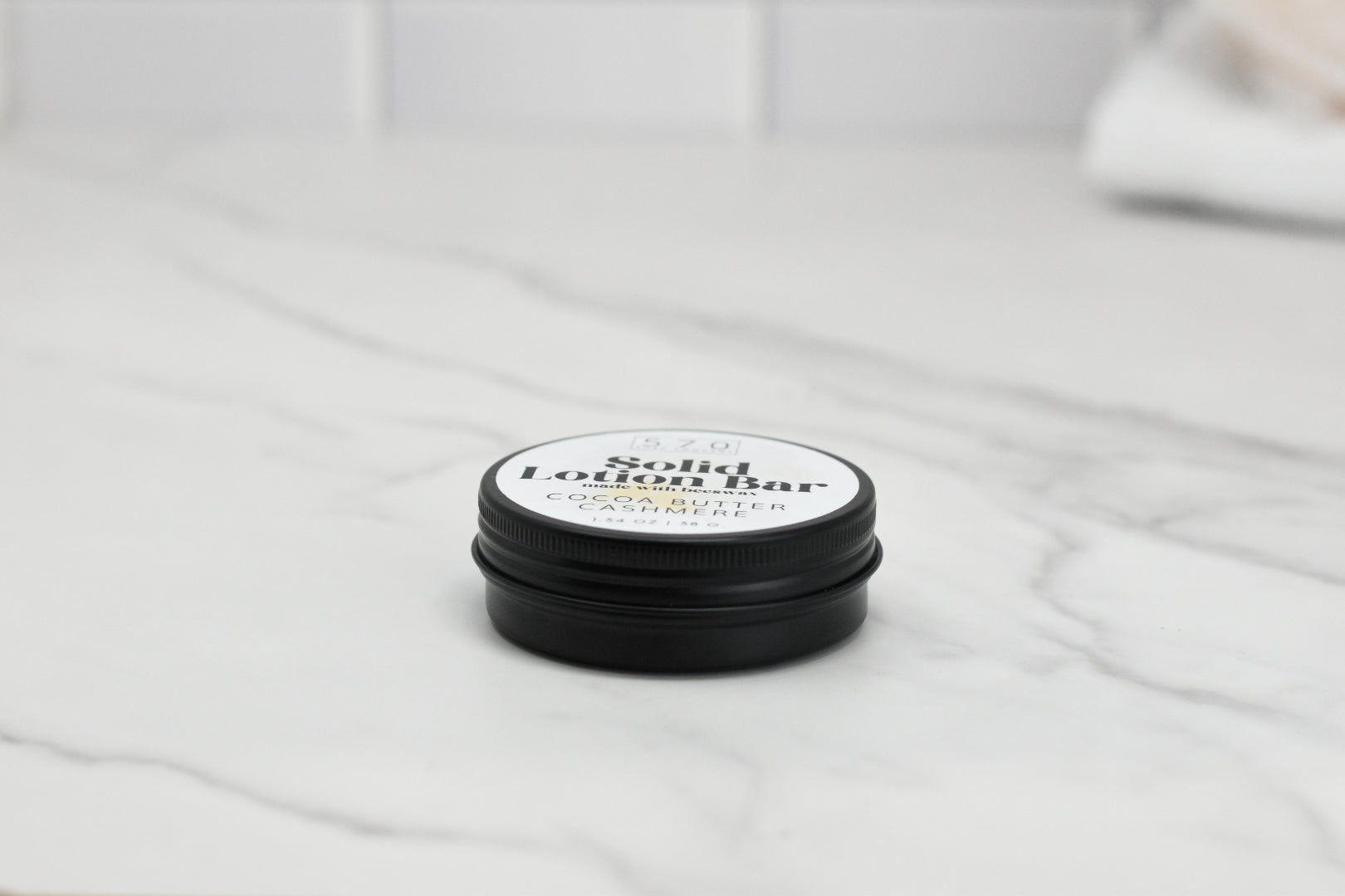 A round black tin labeled "Solid Lotion Bar - made with beeswax" from 570 Soap Co. rests on a marble surface, showcasing its blend of natural ingredients like cocoa butter, shea butter, and cashmere. Priced at $5.20 and perfect as a portable moisturizer, it stands out against the softly blurred background.