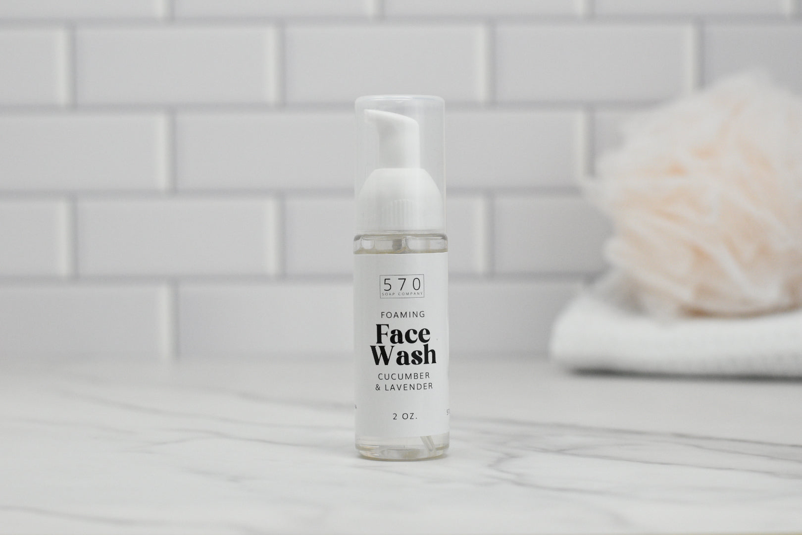 A bottle of Foaming Face Wash - Cucumber Lavender by 570 Soap Co. is placed on a marble surface, infused with soothing Lavender Floral Water and revitalizing Aloe Vera Juice. In the background, a blurred white tile wall highlights a light-colored, fuzzy item resting on neatly folded towels.