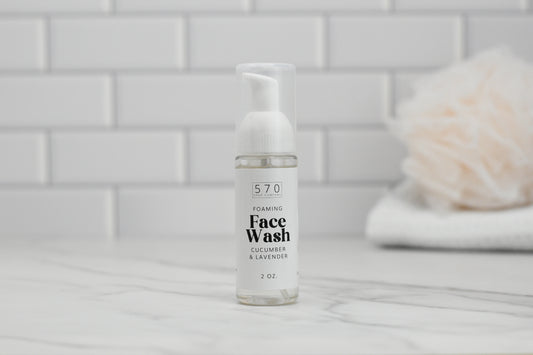 A bottle of Foaming Face Wash - Cucumber Lavender by 570 Soap Co. is placed on a marble surface, infused with soothing Lavender Floral Water and revitalizing Aloe Vera Juice. In the background, a blurred white tile wall highlights a light-colored, fuzzy item resting on neatly folded towels.