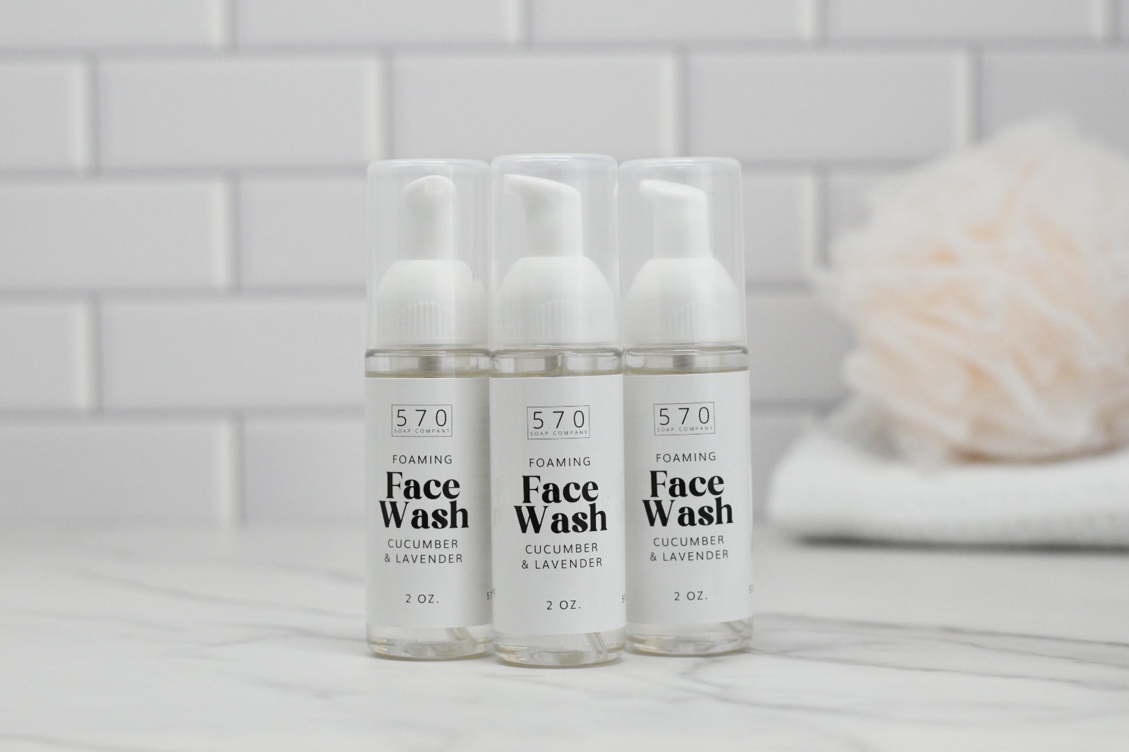 Three 2 oz bottles of the Foaming Face Wash - Cucumber Lavender by 570 Soap Co. are arranged in front of a white tiled wall, complemented by a folded towel in the background. Each bottle promises a refreshing cleanse.