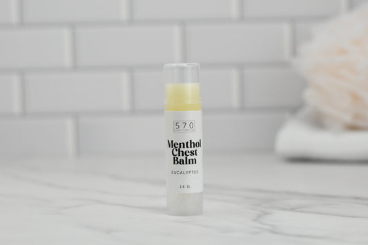 A 14g small tube of 570 Soap Co.'s Menthol Chest Balm - Eucalyptus, featuring natural ingredients and a soothing eucalyptus scent, stands upright on a marble surface. The background showcases a tiled wall and a blurred towel, making it ideal for alleviating respiratory discomfort.